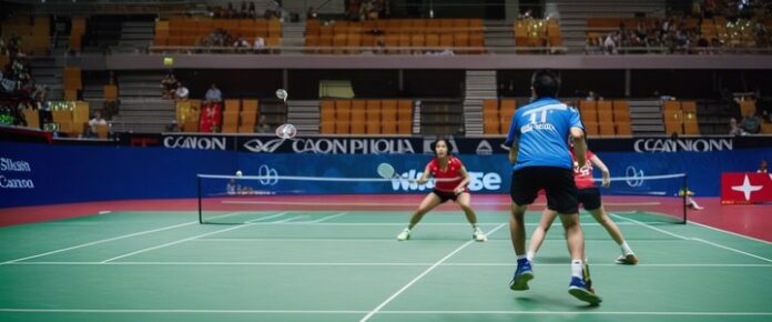 A group of people playing badminton on a green court with a blue and red background.
