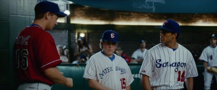 Four baseball players in a dugout, wearing different uniforms and caps, with a crowd of people in the background.